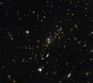 A field of galaxies and with faint curved arcs. Each galaxy appears as a glob of light, most of them reddish-orange. Bright, blue-white, twinkling foreground stars are scattered in a few places in the image.